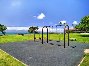 A playground with swings and a bench, located near a grassy area with a view of the ocean. - Tropical Beach Oasis | Private Pool, Hot Tub & AC (Lahaina)