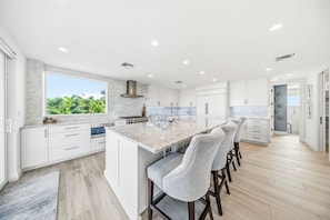 A modern kitchen with a large island, white cabinetry, and a view of greenery outside. - Luxury Home +Carriage house | Waterfront| Pool 5bd (Marathon)