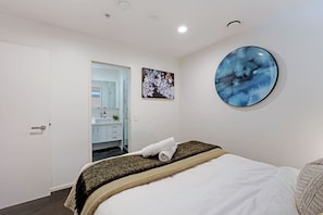 A hotel room with a bed, a round wall art piece, a bathroom visible through an open door, and a small plant on the sink. - Eden Terrace Oasis | Park Views, BBQ & Parking (Auckland Central Suburbs)