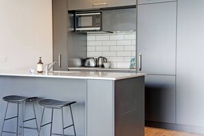 A modern kitchen with a central island, bar stools, a microwave, and a kettle. - Eden Terrace Oasis | Park Views, BBQ & Parking (Auckland Central Suburbs)