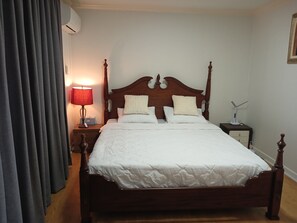 A neatly made bed with a wooden headboard, a bedside lamp, a nightstand, and a window with curtains.