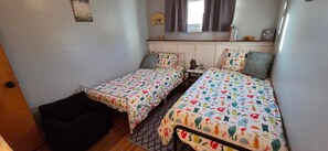 A bedroom with two single beds, a small bedside table, a nightstand, a window with curtains, and a wall-mounted shelf with books and a sign. - 3-bedroom lake house in Hamburg, 10 minutes from Bills Stadium! (Hamburg)