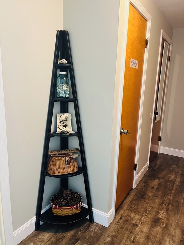 Un coin avec une étagère noire en forme d’échelle, sur laquelle sont disposés des objets décoratifs, une porte en bois et un couloir.