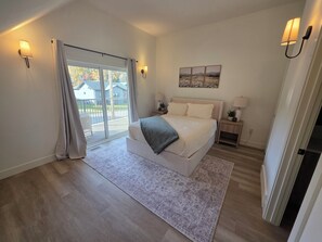 Ein Schlafzimmer mit einem großen Bett, einem Nachttisch, einer Lampe, Blick durch eine Schiebetür und einem Wandbild.