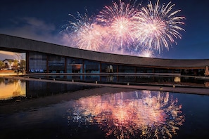 Un edificio con un elemento acuático reflectante al frente, y fuegos artificiales al fondo.