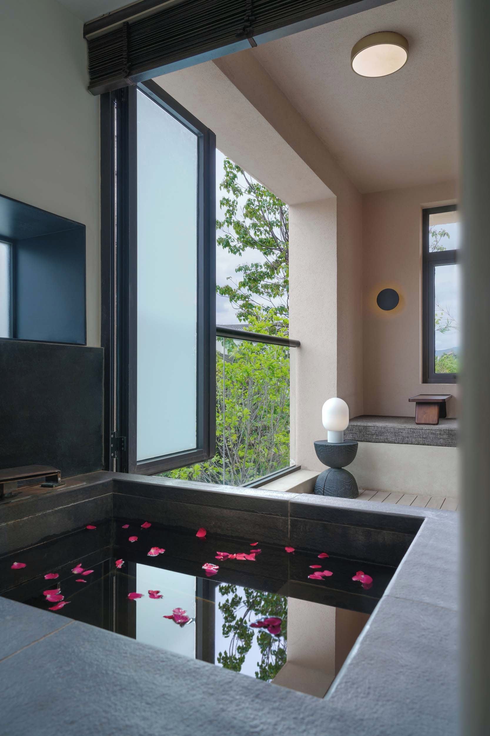 Un baño moderno con un ventanal grande, una bañera con pétalos de rosa, una lámpara de techo redonda y un televisor de pantalla plana.