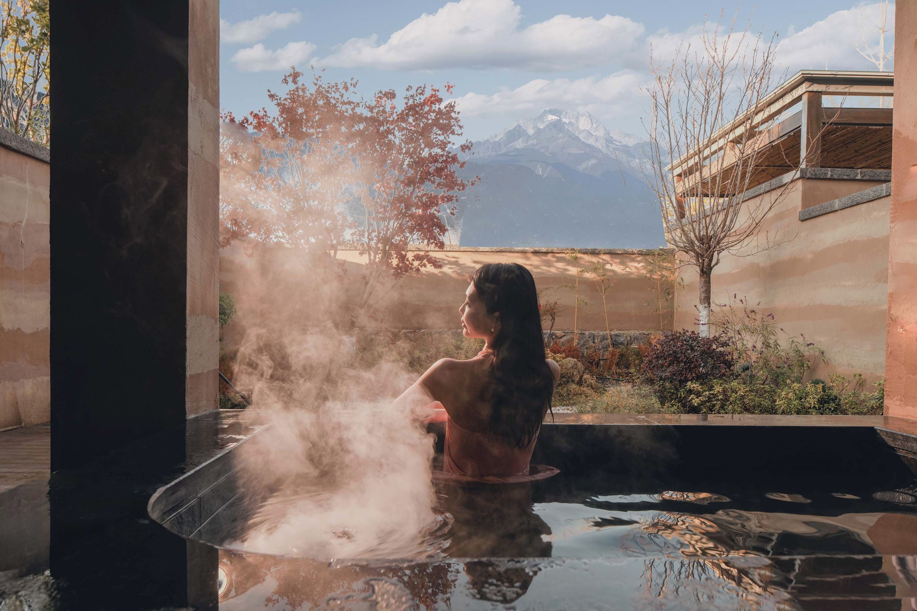 A person in a hot tub with steam, overlooking a mountainous landscape and a building with a balcony.