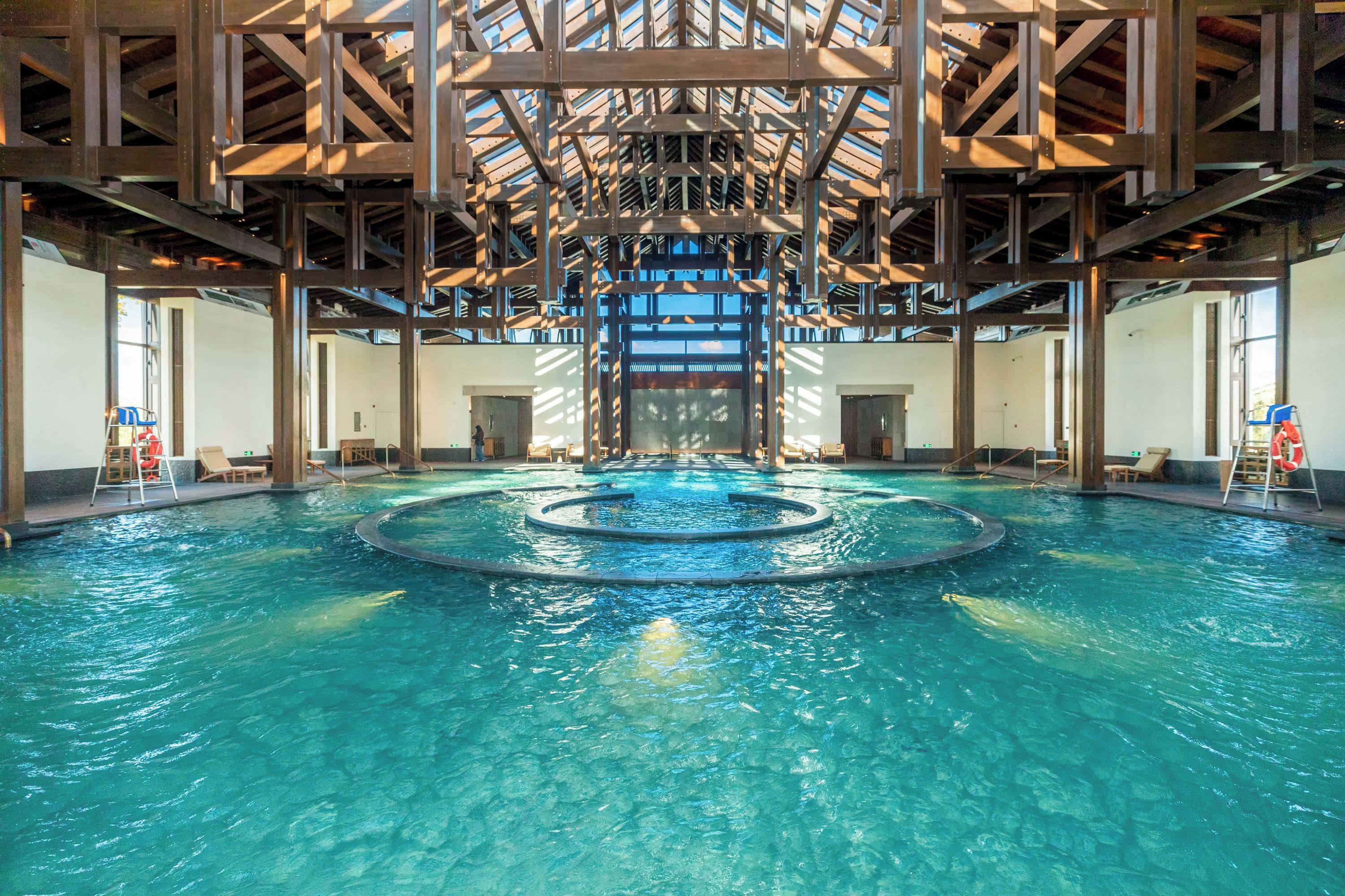 Una piscina interior con una fuente central, rodeada de áreas de descanso y un techo alto con vigas a la vista.
