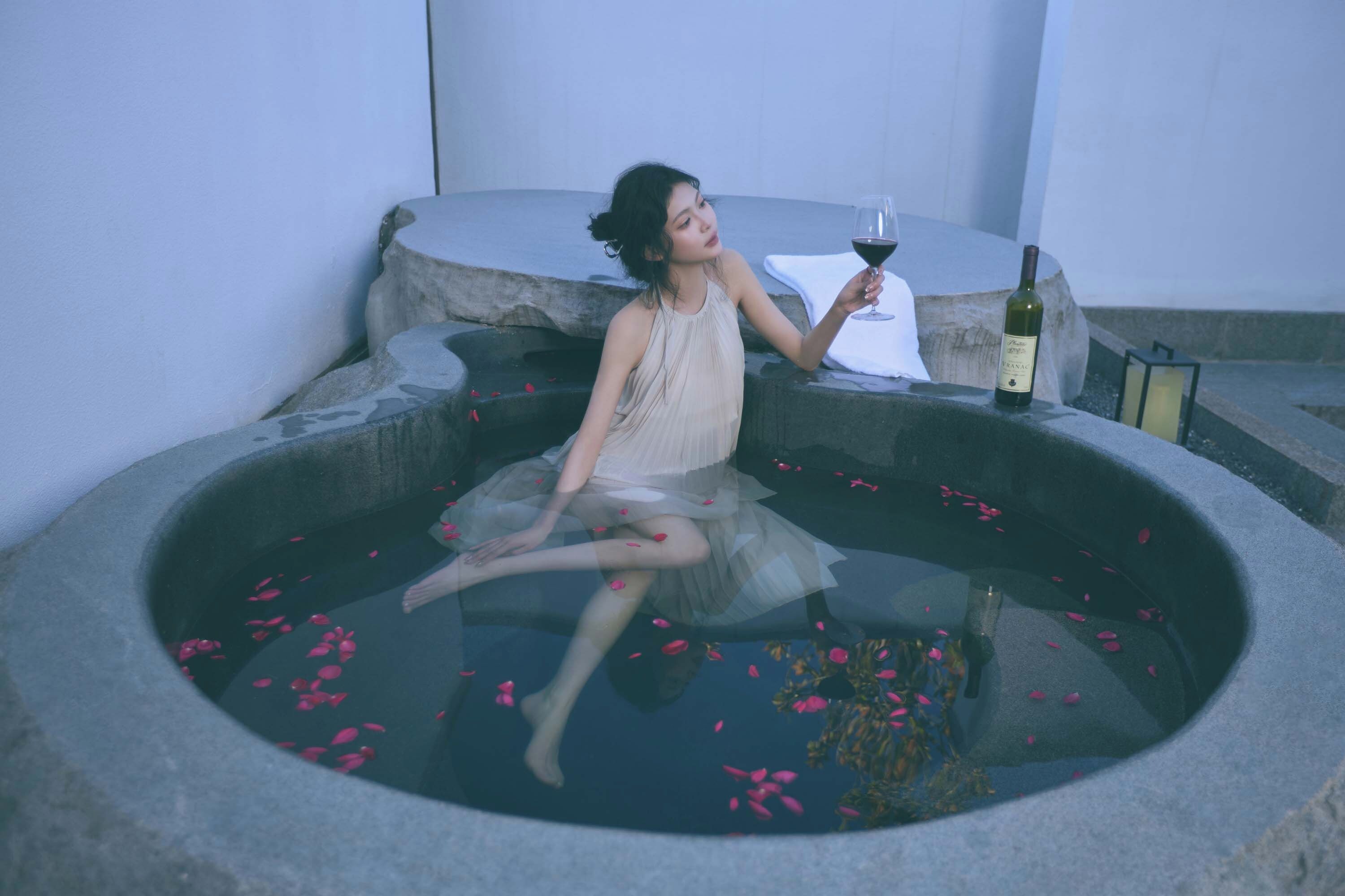 Pessoa sentada em banheira de pedra com pétalas de rosa, segurando uma taça de vinho e uma garrafa de vinho na borda.