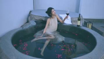 Pessoa sentada em banheira de pedra com pétalas de rosa, segurando uma taça de vinho e uma garrafa de vinho na borda.