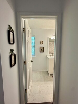 A hallway leading to a bathroom with a white door, a framed picture on the wall, and a window with blinds. -  Stylish 3BR Home with Fireplace (Marietta)