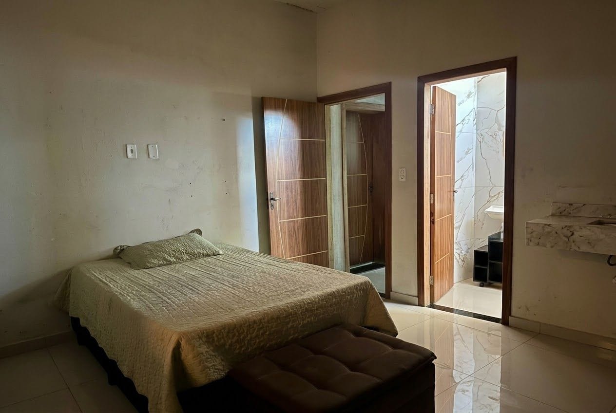 Quarto com cama, um banquinho, guarda-roupa e banheiro visível por uma porta aberta.