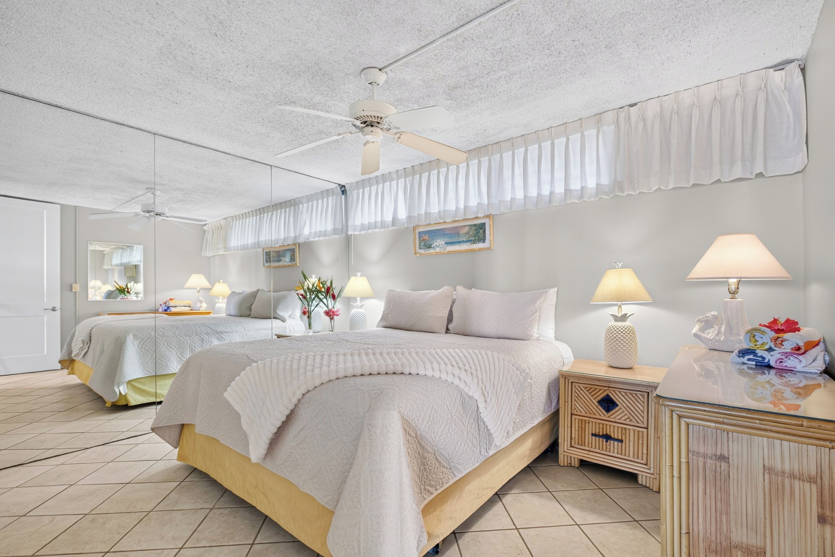 A hotel room with a large bed, two bedside tables, a ceiling fan, and a view of the sea through the window.