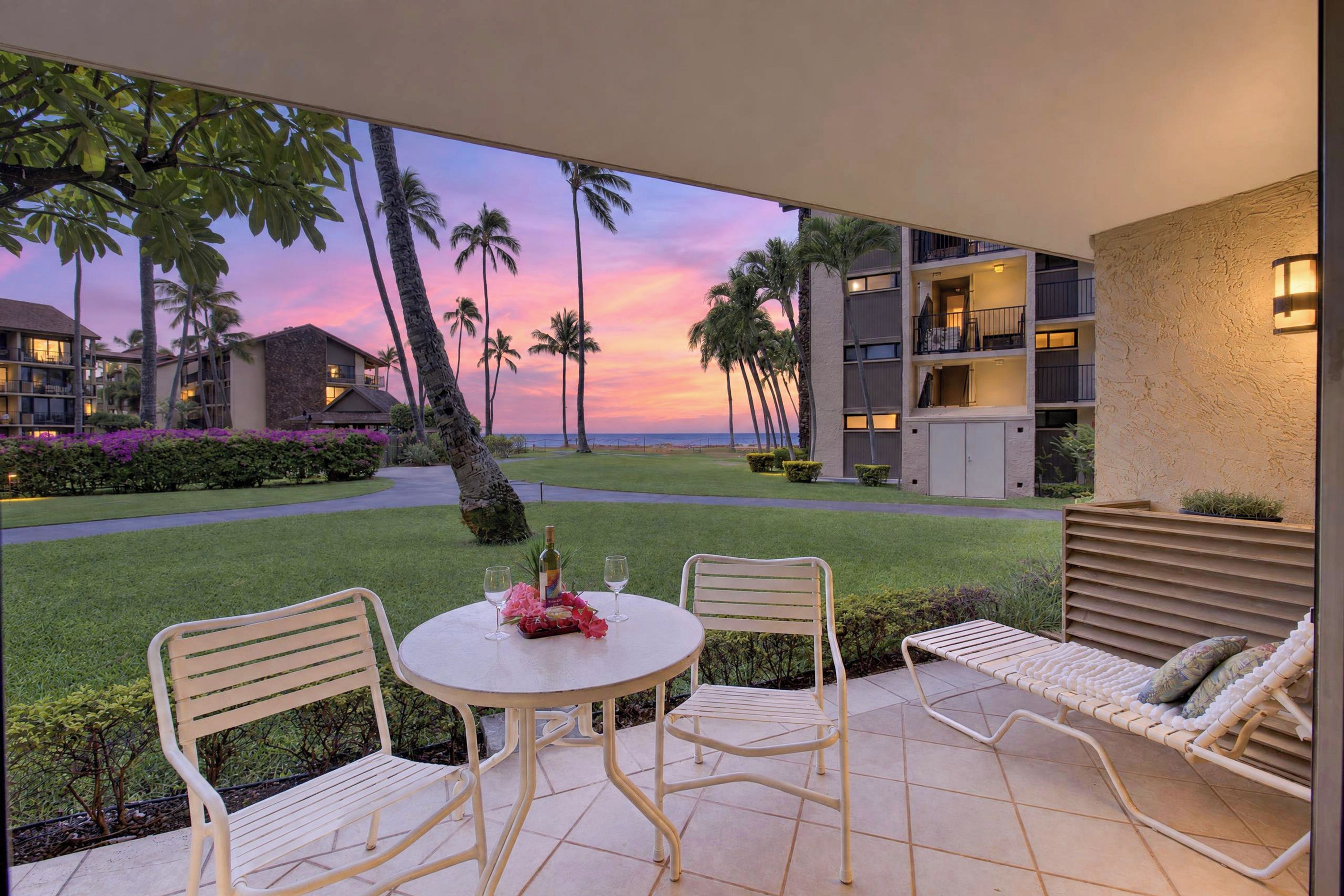 A patio with two chairs and a table, overlooking a landscaped area with palm trees and a building with balconies.