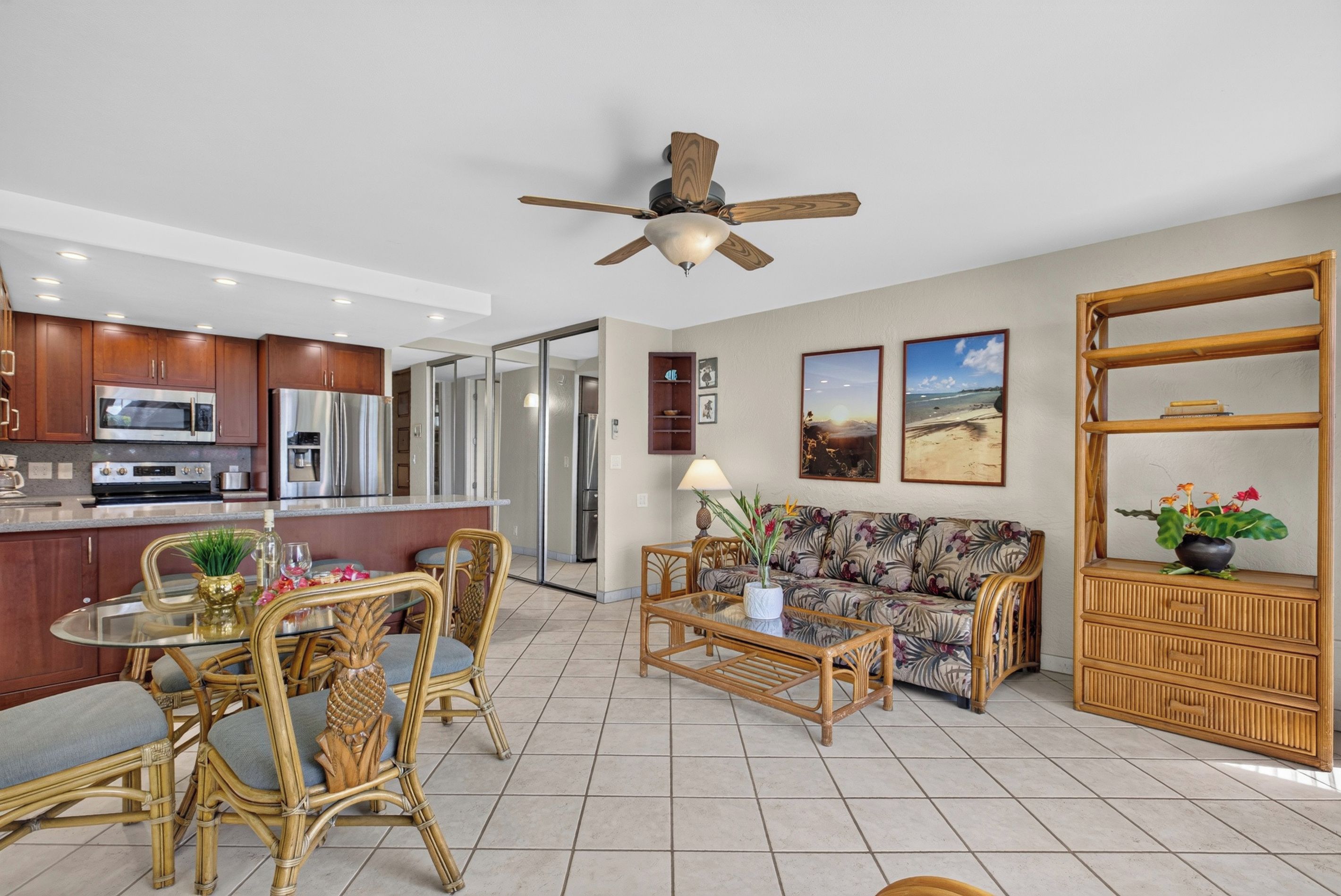 A modern kitchen with stainless steel appliances, a dining area with wicker furniture, and a living room with a sofa and a ceiling fan.