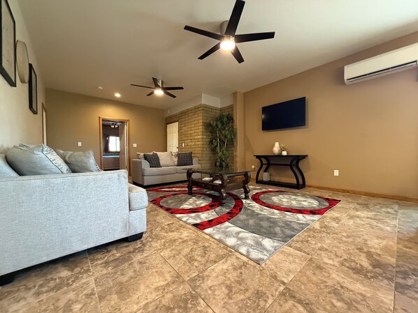 A living room with a grey sofa, a coffee table, a red and grey rug, a ceiling fan, and a television mounted on the wall.