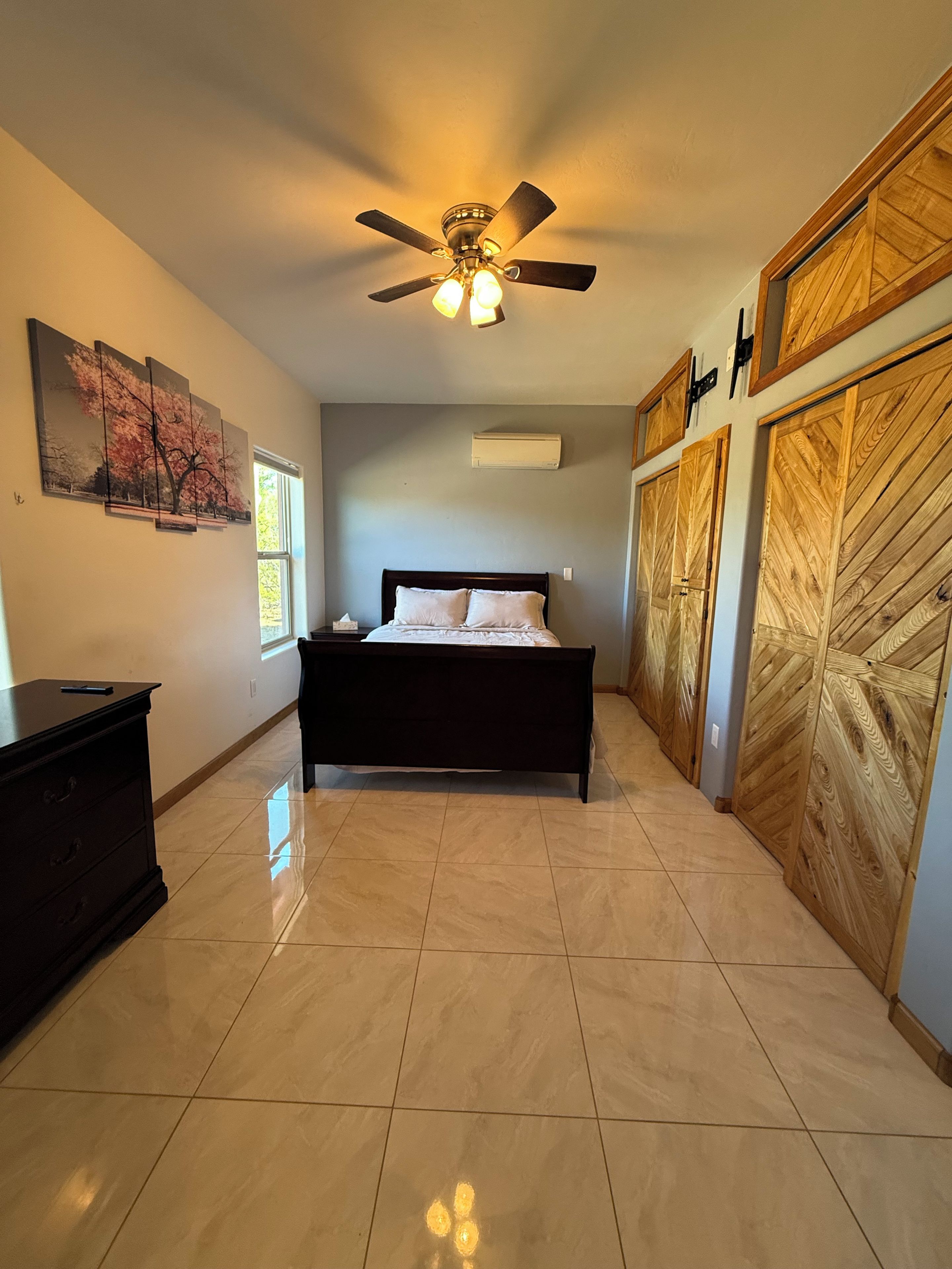 Une chambre à coucher avec un lit, un ventilateur de plafond, des œuvres d’art et une armoire en bois.