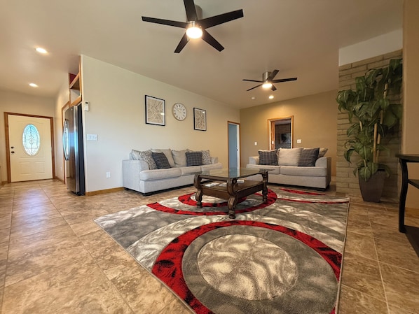 A spacious living room with two sofas, a coffee table, and a large rug. There are ceiling fans, a door with a glass panel, and a potted plant.