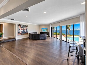 A spacious living area with a large window overlooking a pool and ocean view, a dark-colored sectional sofa, and a flat-screen TV. - Bayfront 6BR Compound | Pool, Hot Tub & Views (Miami)