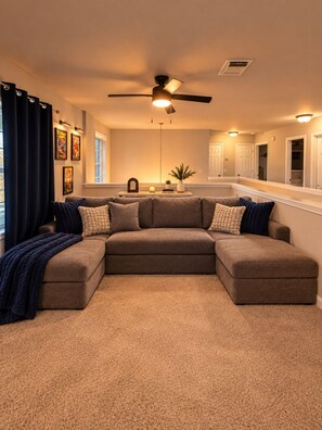 A living room with a sectional sofa, a ceiling fan, and a small table with decorative items. - Cedar Park Oasis - Pool, Movie & Game Room, BBQ, Sleeps 18 (Cedar Park)