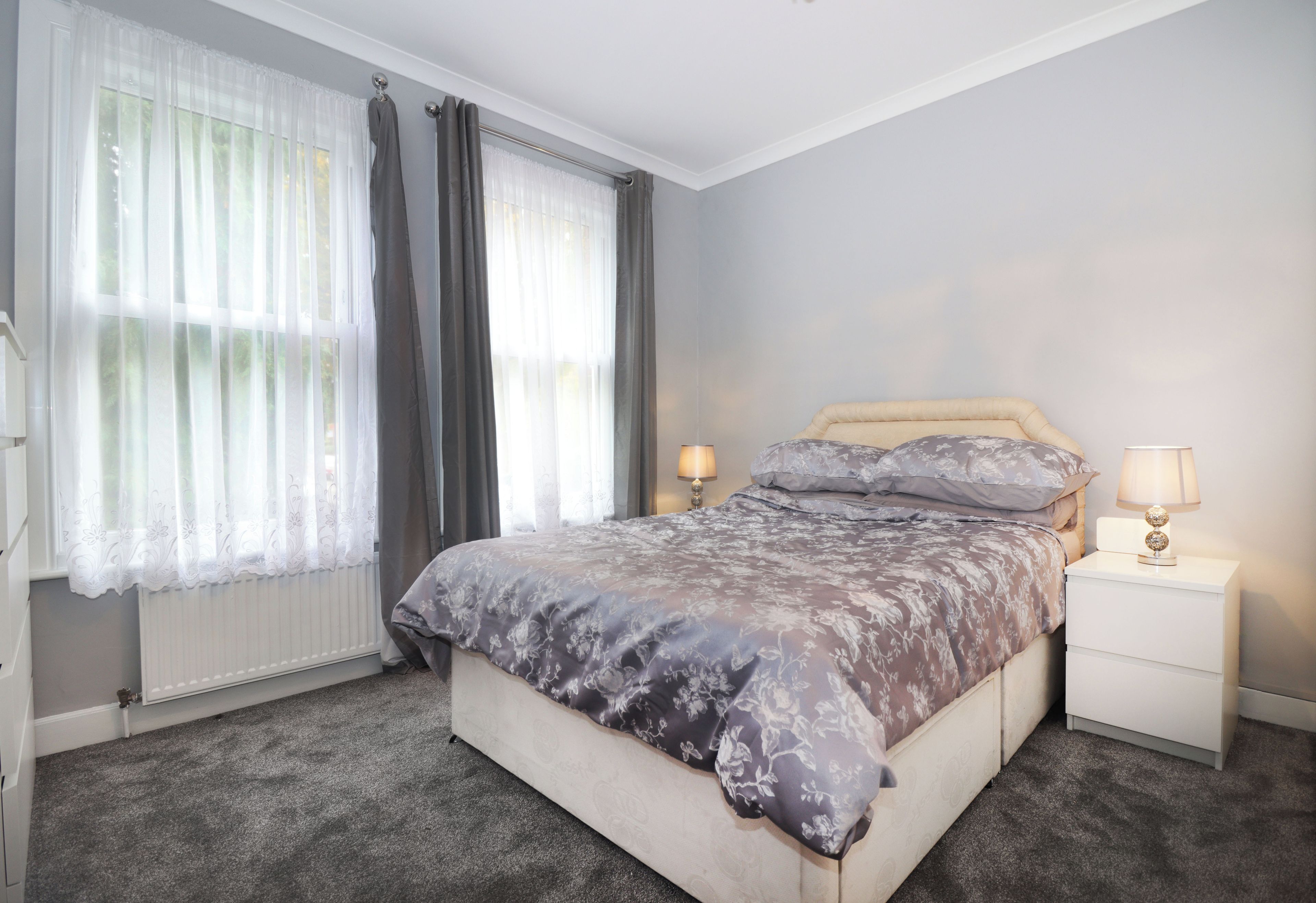 A bedroom with a bed, two bedside tables, a window with curtains, and a radiator.