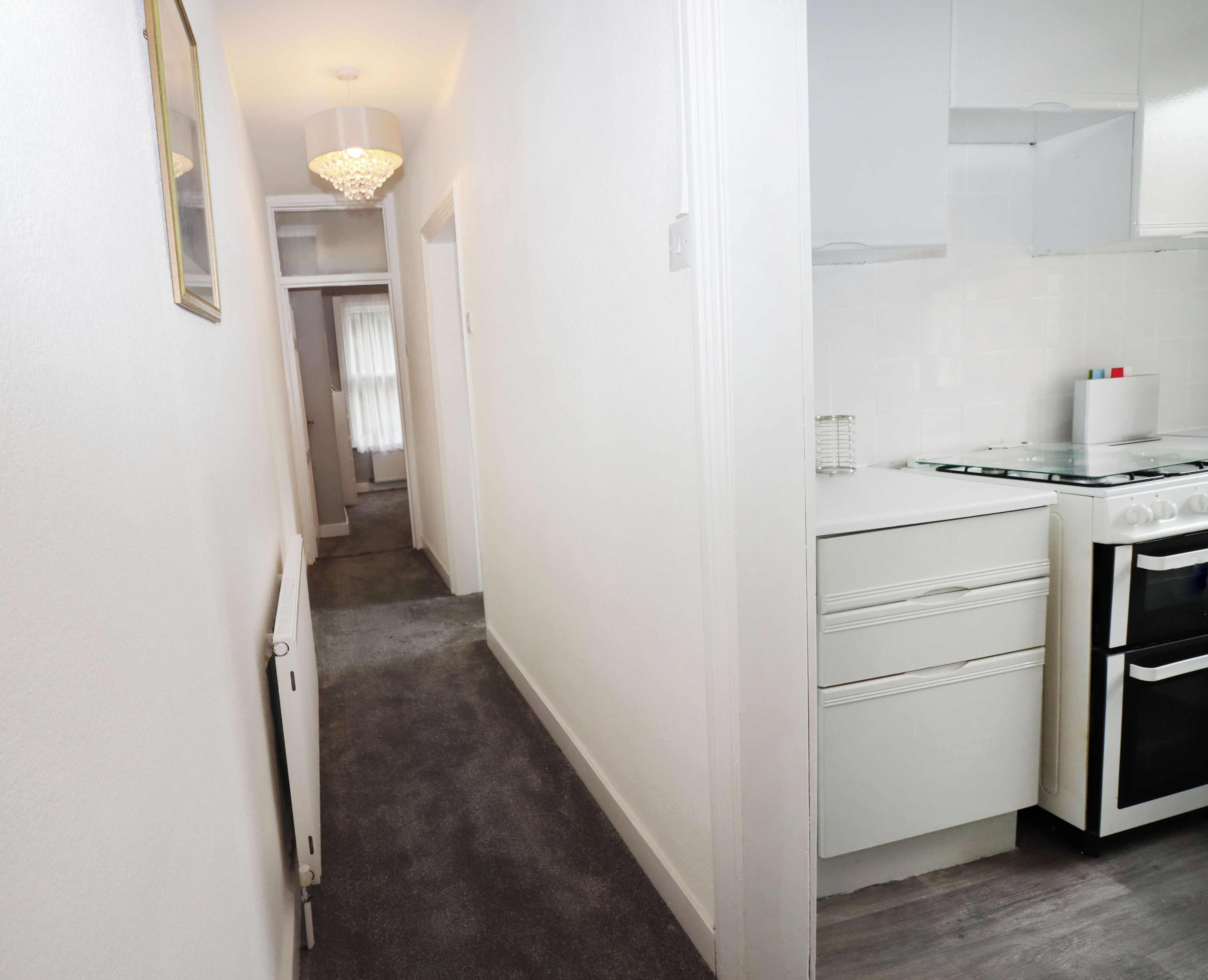 A narrow hallway leading to a kitchen with white cabinets, a glass-top table, and a black oven.