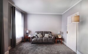Ein Wohnzimmer mit einem grauen Sofa, einem kleinen Tisch mit einer Lampe und einer Tür.