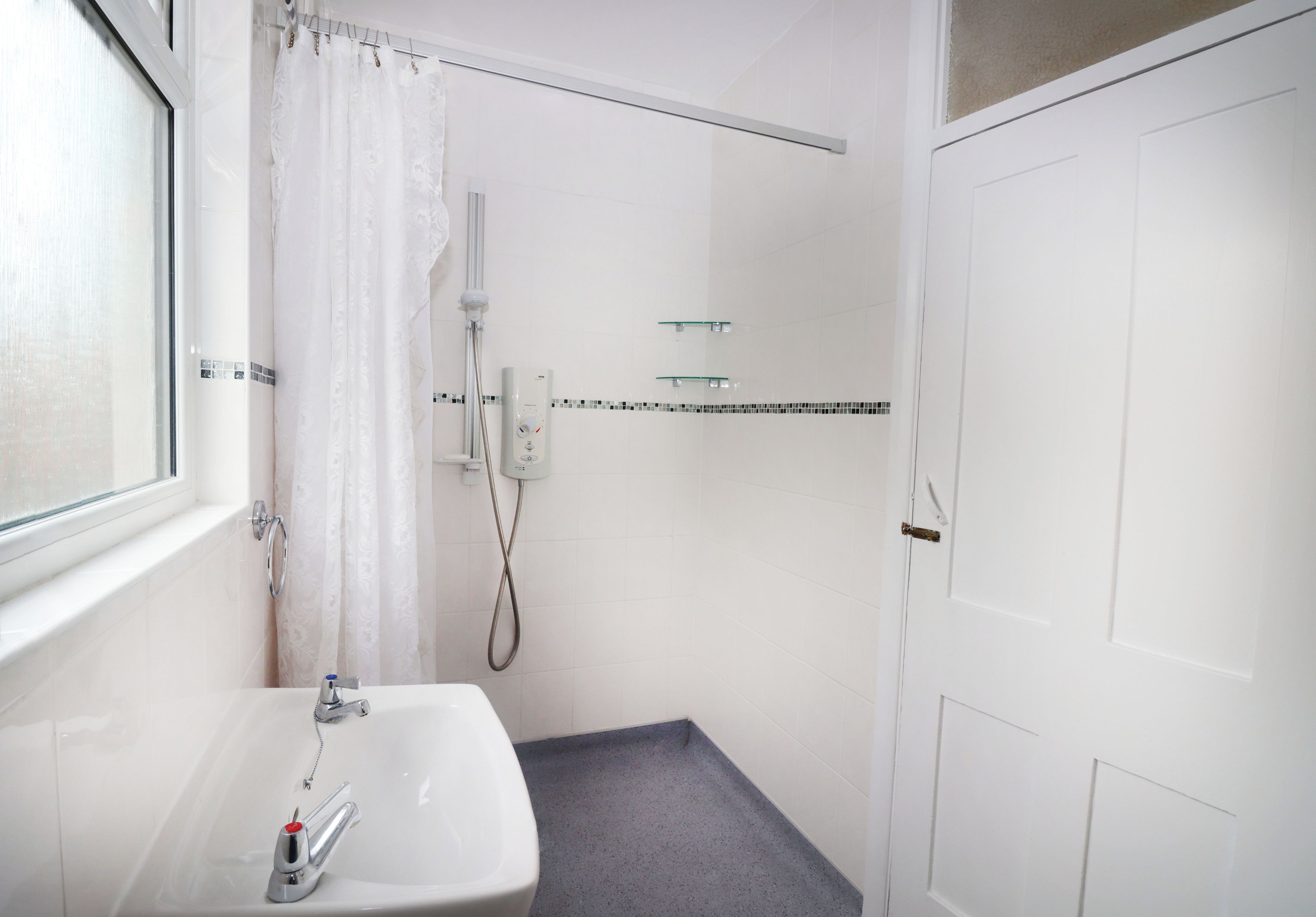 A bathroom with a white sink, a shower with a glass shelf, and a white door.