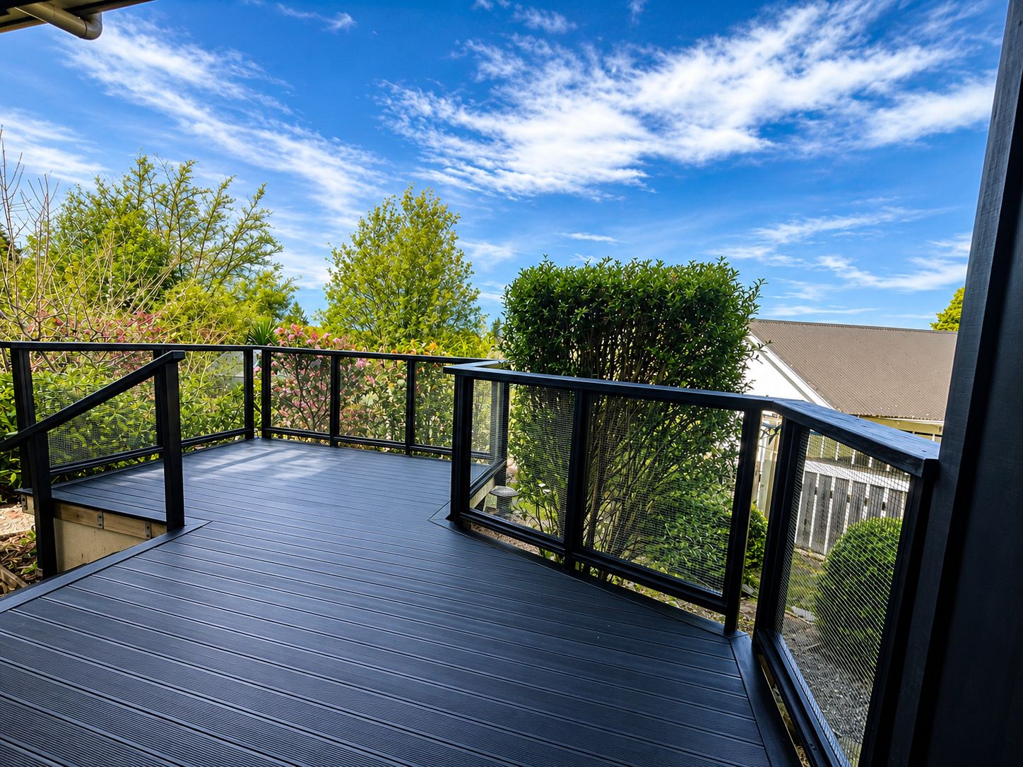 A wooden deck with a black railing, surrounded by greenery and trees under a clear sky.