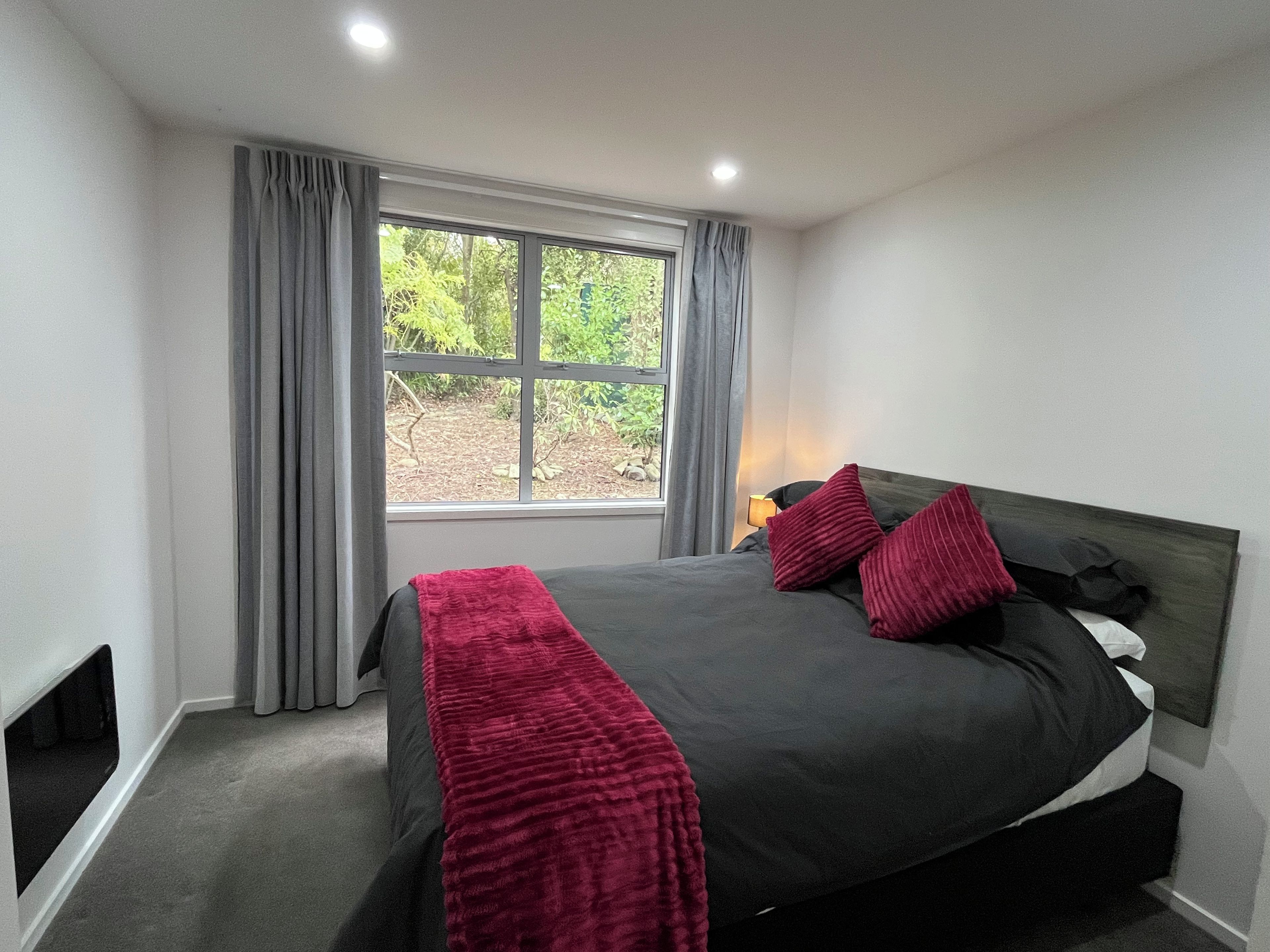 Ein Schlafzimmer mit einem großen Bett, grauen Vorhängen, einem Fenster mit Blick auf Grünflächen und einer Nachttischlampe.
