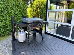 Un barbecue portatif avec une bouteille de propane, placé sur une terrasse d’où l’on peut admirer une maison et la verdure environnante.