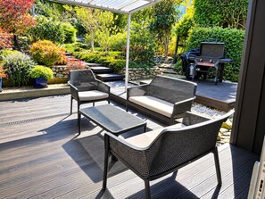 Une terrasse extérieure aménagée avec des meubles en osier, un barbecue et un jardin où poussent différentes espèces de plantes.