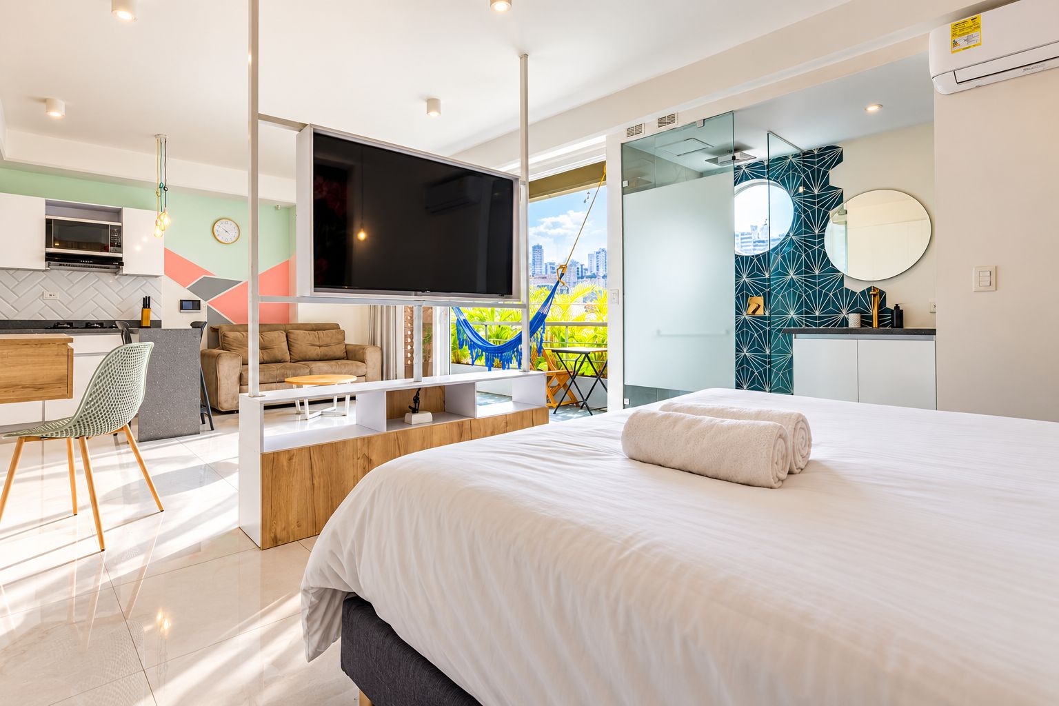 A modern hotel room with a large bed, a flat-screen TV, a dining area, and a view of the city.