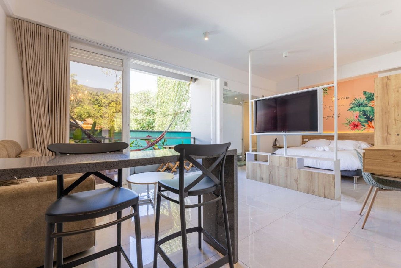 Habitación de hotel moderna con zona de comedor, cama con televisor y ventana grande con vistas a zonas verdes.