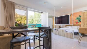 Habitación de hotel moderna con zona de comedor, cama con televisor y ventana grande con vistas a zonas verdes.