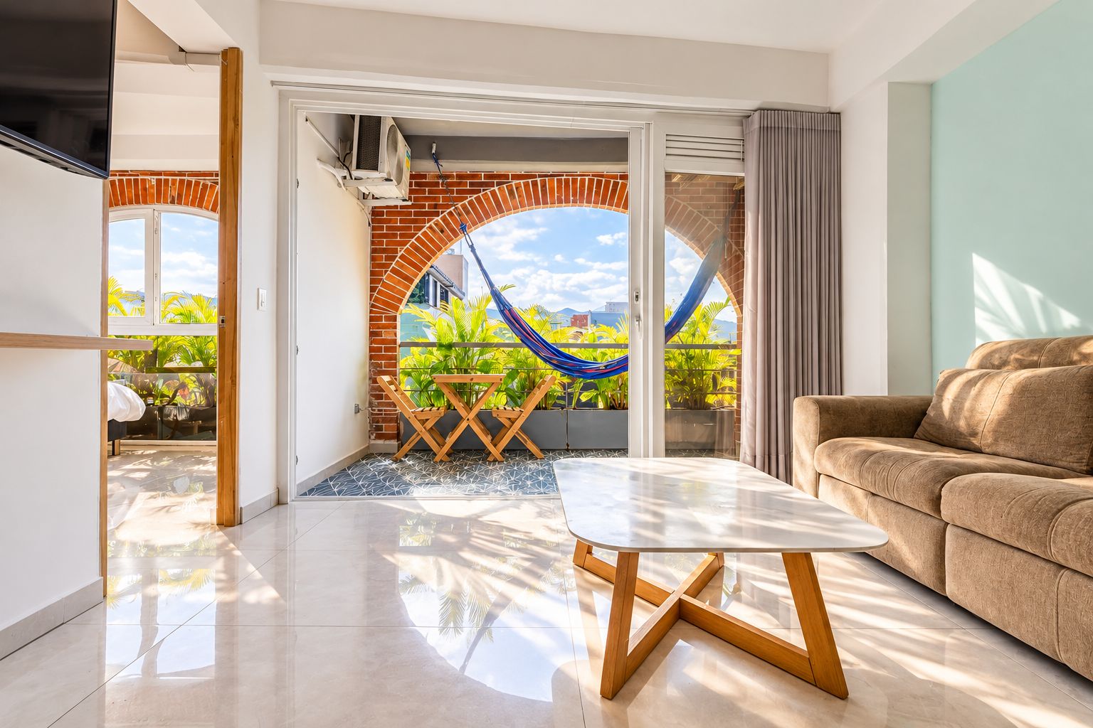 A modern living room with a sofa, a dining table with chairs, and a view of a balcony with a hammock.