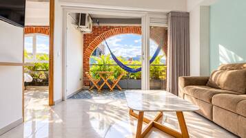 A modern living room with a sofa, a dining table with chairs, and a view of a balcony with a hammock.