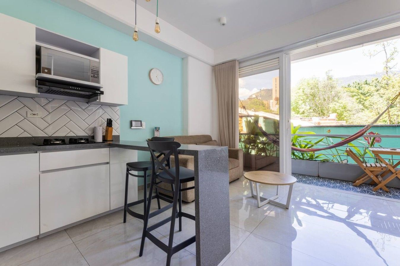 Una cocina moderna con salpicadero de azulejos tipo espiga, microondas integrado y cocina a gas. Hay una zona de estar con sofá y una mesa redonda pequeña, y un balcón con una hamaca.