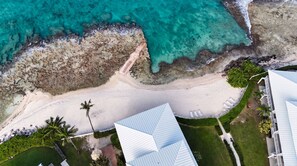 An aerial view of a coastal property with a beach, palm trees, and a building with a white roof. - New Re-Furnished 2bd Condo with Ocean Views from Every Room on Seven Mile Beach (George Town)