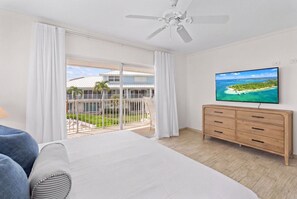 A hotel room with a bed, a TV, a balcony with a view of a beach, and a wooden dresser. - New Re-Furnished 2bd Condo with Ocean Views from Every Room on Seven Mile Beach (George Town)
