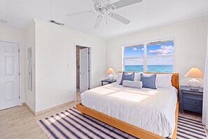 Quarto com cama, ventilador de teto, janela vista para o mar e tapete listrado.