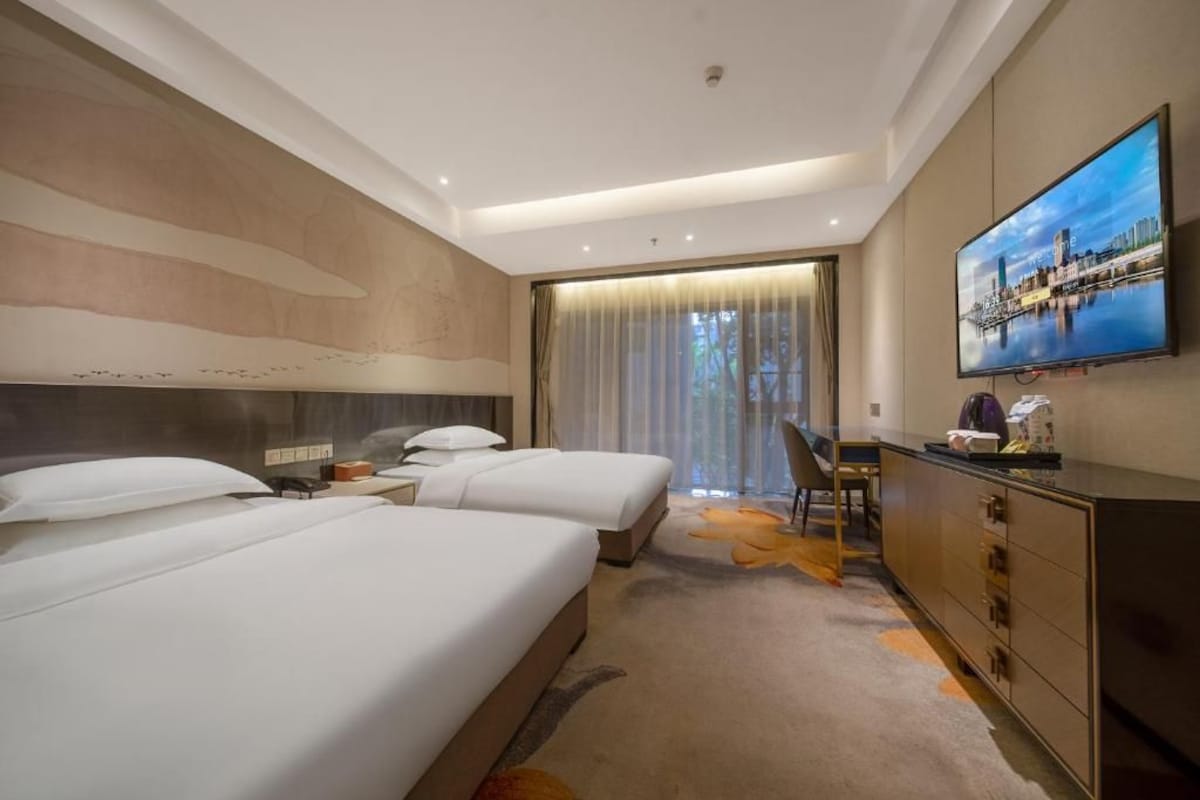 Ein Hotelzimmer mit zwei Betten, einem Schreibtisch, einem Fernseher und Blick auf eine Stadtansicht.