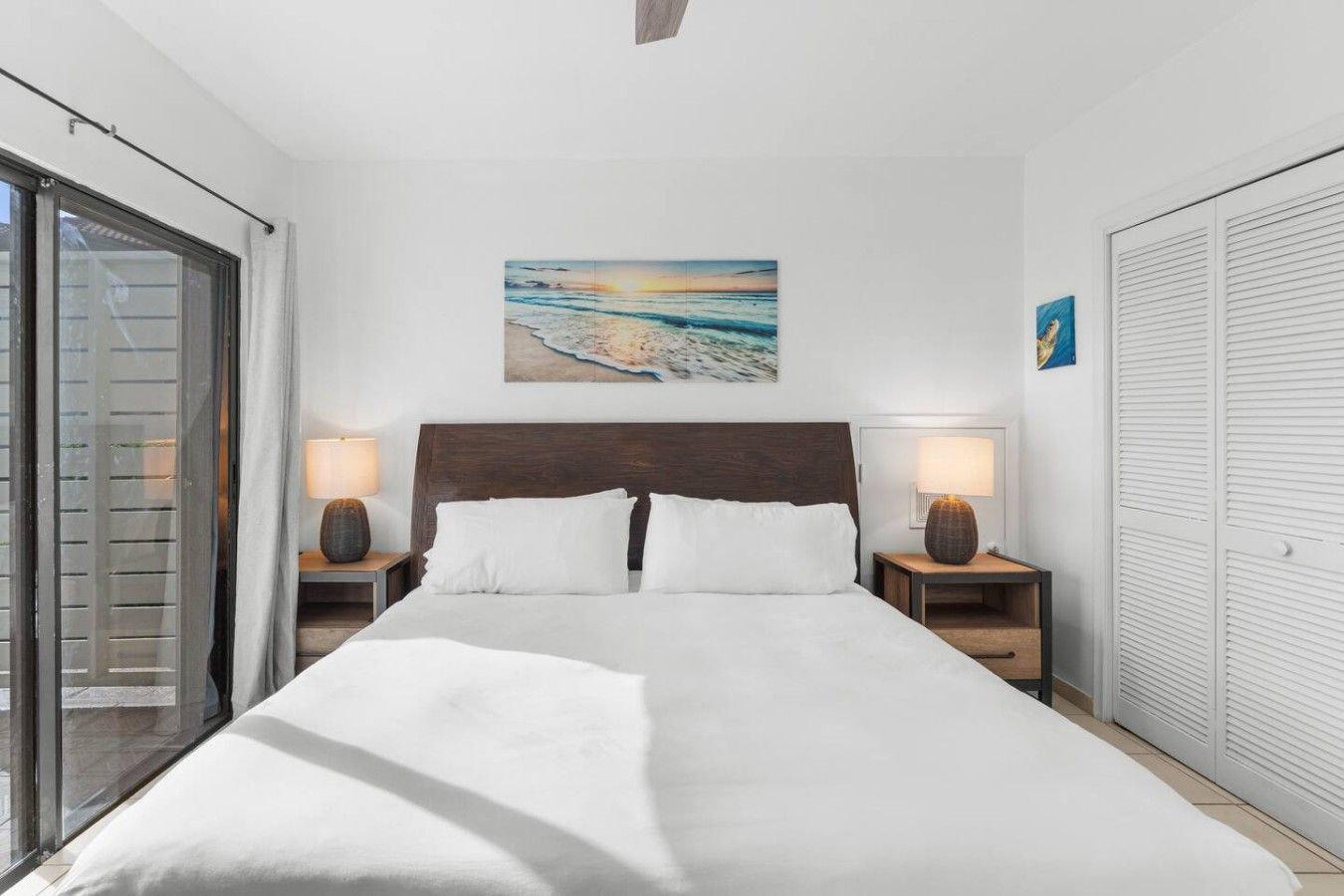 Una habitación de hotel con una cama grande, dos mesitas de noche con lámparas, un cuadro en la pared y un ventanal corredero.