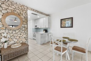 Una cuina moderna amb armaris blancs, una paret d'accent de pedra, una zona de menjador amb taula de vidre i cadires blanques, i un ventilador de sostre.