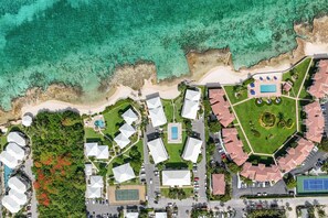 An aerial view of a resort with multiple buildings, a swimming pool, tennis court, and a beach. - Newly Renovated Beachfront 2BR w Ocean & Pool View (George Town)