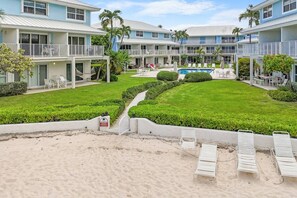 A beachfront hotel with multiple-story buildings, a swimming pool, and a sandy beach area with lounge chairs. - Newly Renovated Beachfront 2BR w Ocean & Pool View (George Town)