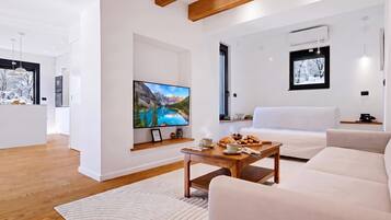 A modern living room with a flat-screen TV, a wooden coffee table with breakfast items, a white sofa, and a large window with a mountain view.