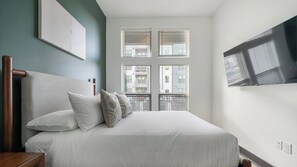 Ein Schlafzimmer mit einem Bett, Kissen, einem Nachttisch, einem an der Wand befestigten Fernseher und einem Fenster mit Jalousien.
