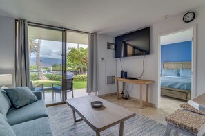 A modern hotel room with a sofa, a dining table, a TV mounted on the wall, and a bed with a blue headboard. - Oceanfront Kihei Condo | Ground Floor, Pool, Hot Tub & AC (Kihei)
