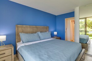 A bedroom with a large bed, a wooden headboard, a bedside table with a lamp, and a view of a garden through an open door. - Oceanfront Kihei Condo | Ground Floor, Pool, Hot Tub & AC (Kihei)